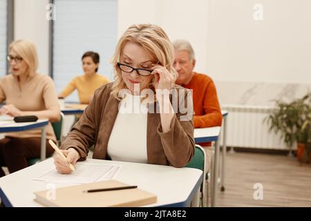 Ernst reife Frau in Brillen sitzen am Schreibtisch im Klassenzimmer und schriftlich Test mit Menschen im Hintergrund Stockfoto