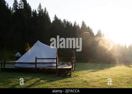 Seitenansicht des weißen Glamping-Zeltes auf einer Holzterrasse auf einer grünen Wiese, umgeben von Tannenwäldern bei Sonnenaufgang Stockfoto