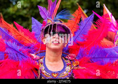 Notting Hill, London, Großbritannien. 29. August 2022. Europas größtes Straßenfest ist nach der Annullierung der Jahre aufgrund der Covid-Pandemie auf die Straßen von Notting Hill zurückgekehrt. Jamaikanische, exotische Tänzer und Musikgruppen zogen durch die Straßen, wobei Essen und Straßenunterhaltung in der Umgebung zur Veranstaltung hinzufügten. Die Grand Parade findet am Montag der Bankfeiertage statt, als Höhepunkt des dreitägigen Festivals, das 1966 begann. Weiße kaukasische Frau trägt ein farbenprächtig gefärbtes Federkostüm Stockfoto