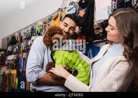 Lächelndes interrassisches Paar mit Tierjacke auf Pudel im Tierladen Stockfoto