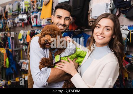 Lächelndes, multiethnisches Paar, das eine Tierjacke auf Pudel trägt und im Tierladen die Kamera anschaut Stockfoto