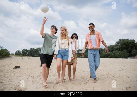 Junger Mann spielt mit Ball, während er mit multiethnischen Freunden am Sandstrand spazieren geht Stockfoto