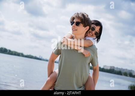 Junger Mann in Sonnenbrille Huckepack fröhlich asiatische Frau in der Nähe von Fluss Stockfoto