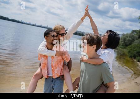 Interracial Männer Huckepack Frauen geben hohe fünf am Flussufer Stockfoto