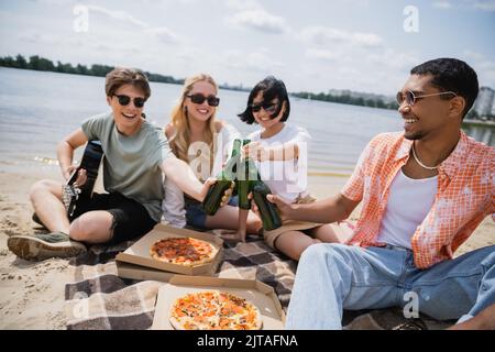 Glückliche interrassische Freunde in Sonnenbrillen klirrenden Bierflaschen während der Strandparty Stockfoto