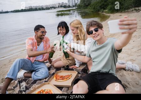 Lächelnder Mann beim Selfie mit multiethnischen Freunden, die in der Nähe der Pizza auf Bierflaschen klimmten Stockfoto