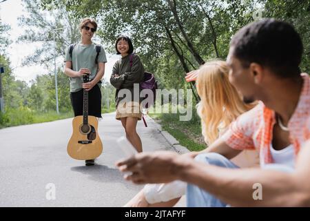 Lächelnder Mann mit Gitarre in der Nähe einer asiatischen Frau und verschwommene interrassische Freunde, die auf der Straße sitzen Stockfoto