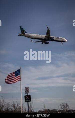 Eine niedrige Ansicht eines Jet-blauen Flugzeugs im Flug, das über einer amerikanischen Flagge landet Stockfoto