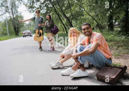 Verschwommene Anhalter halten das Auto in der Nähe lächelnder interrassischer Freunde, die auf der Straße sitzen Stockfoto