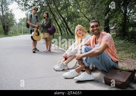 Lächelnde, interracial Reisende, die auf der Straße in der Nähe verschwommener Freunde mit Gitarre sitzen Stockfoto