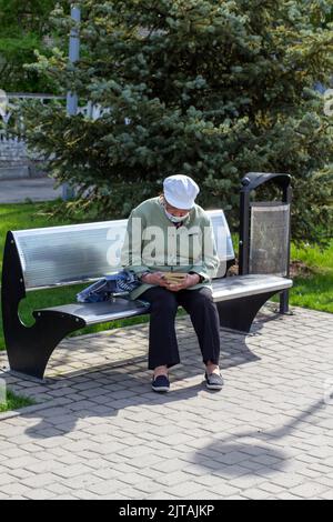 Novomoskovsk, Ukraine - 13. Mai 2021: Ältere Frau in Maske auf Bank auf der Straße in der Stadt liest Buch Stockfoto