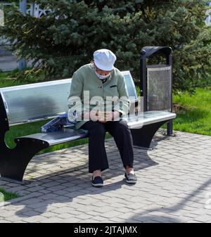 Novomoskovsk, Ukraine - 13. Mai 2021: Ältere Frau in Maske auf Bank auf der Straße in der Stadt liest Buch Stockfoto