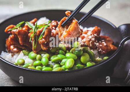 Teriyaki-Huhn mit Reis, Zwiebeln und Edamame-Bohnen in einer schwarzen Schüssel, Nahaufnahme. Japanische Küche. Stockfoto