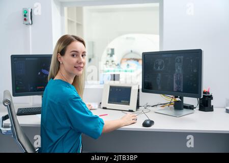 Frau in medizinischer Kleidung sitzt halb gedreht Tisch und schaut in die Linse Stockfoto