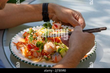 Hausgemachter Papaya-Salat mit Meeresfrüchten, Garnelen und Lachs auf einem weißen Teller mit männlichen Händen und Essstäbchen, draußen in Sonne und Schatten Stockfoto