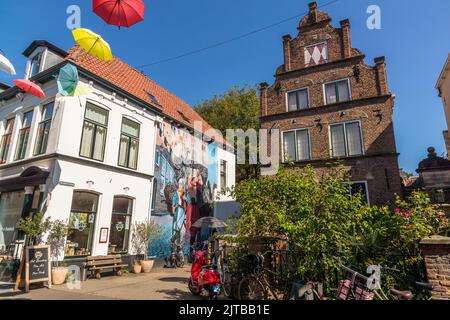 Ein Wandgemälde, das das Hansische Jahr 2023 markiert – es zeigt eine Marktfrau, die Deventer Honigkuchen und Stockfisch verkauft, wichtige Waren des Hanshandels, zusammen mit ihrem Sohn, und verbindet die Vergangenheit und Gegenwart der Stadt – schmückt die Giebelwand eines Hauses in der Altstadt von Deventer, einer von neun historischen Hansestädten an der IJssel, Provinz Overijssel, Niederlande. Der Junge im Wandgemälde trägt ein Fanhalsband des Fußballvereins Deventer Eagles. Bunte Sonnenschirme hängen über der Straße davor, und ein rotes Moped sowie mehrere Fahrräder sind am Haus geparkt. Stockfoto