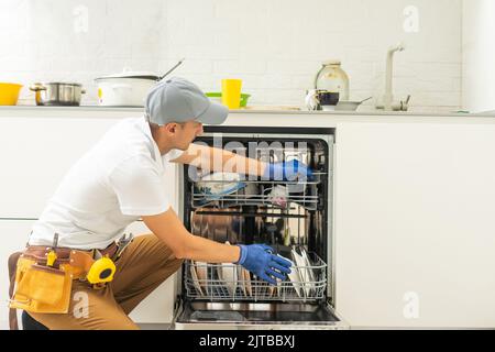 Reparaturman in Uniform Reparaturen Geschirrspüler in der Küche. Der junge Mann-Spezialist entschrauben Teile mit einem Schraubendreher, um den Zustand der Tabletts auf die Seite der Schale zu überprüfen. Stockfoto