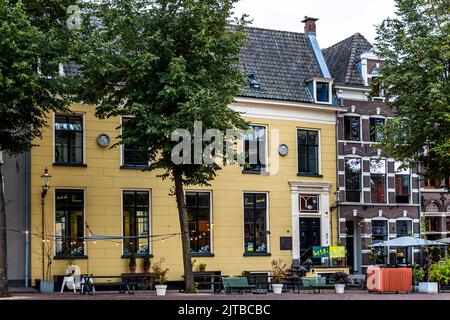 Ein gelb gestrichenes Gebäude mit der Aufschrift „Hotel“ steht an einer von Bäumen gesäumten Straße in Deventer, einer von neun historischen Hansestädten an der IJssel, Provinz Overijssel, Niederlande. Ein Außensitzbereich mit Tischen und Bänken, die mit Lichterketten geschmückt sind, lädt Passanten ein, vor dem Gebäude zu verweilen, wobei rechts ein Giebelhaus und ein rot-weißer Sonnenschirm angrenzen. Stockfoto