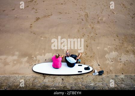 Blick auf ein älteres Paar, das auf einem Surfbrett an einem Sandstrand sitzt Stockfoto