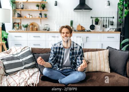 Ruhiger, glücklicher, friedlicher, entspannter kaukasischer junger Mann in lässiger Kleidung, allein zu Hause im Wohnzimmer auf der Couch sitzend und lächelnd in der Lotusposition meditierend. Entspannungs- und Meditationskonzept Stockfoto