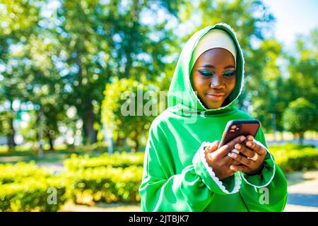 lateinamerikanische hispanic arabische Frau in grünem muslimischen Kleid mit modernem hellen Make-up und Nasenpiercing Blick auf Telefonbildschirm im Freien Stockfoto