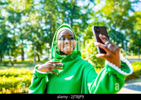 Multikulturelle Woman in grünem muslimischen Kleid mit hellem Make-up und durchdringender Nase, die im Sommer Selfie im Freien macht Stockfoto