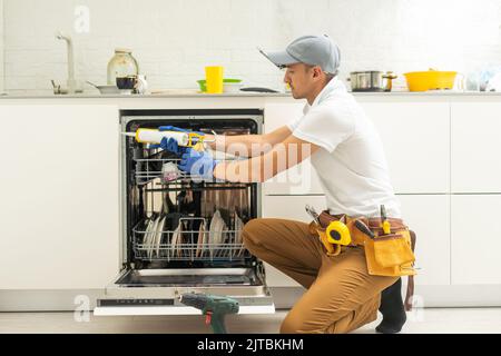 Reparaturman in Uniform Reparaturen Geschirrspüler in der Küche. Der junge Mann-Spezialist entschrauben Teile mit einem Schraubendreher, um den Zustand der Tabletts auf die Seite der Schale zu überprüfen. Stockfoto