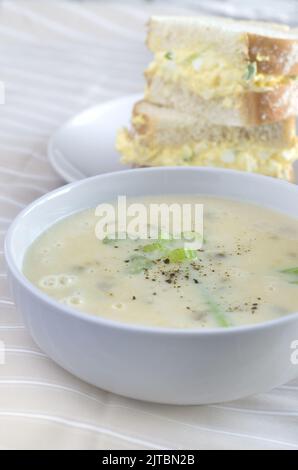Creme der Sellerie-Suppe in einer weißen Schüssel mit verschwommenem Eiersalat-Sandwich im Hintergrund vertikal Stockfoto