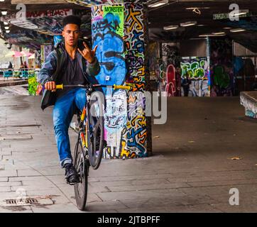 Ein schwarzer Jugendlicher, der mit einem Rad auf seinem Fahrrad fährt, macht im Undercroft Skate Park in South Bank, London, England, UK mit den Fingern eine Geste zur Kamera Stockfoto