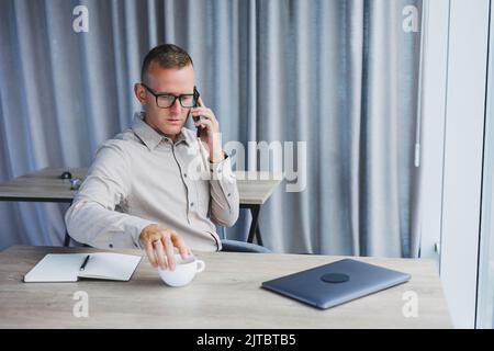 Innenaufnahme des fröhlichen bärtigen männlichen Freiberuflers Arbeit in fernen, verwaltet Finanzen über Smartphone, sitzt am Holztisch, arbeitet mit Dokumentation, hat Stockfoto