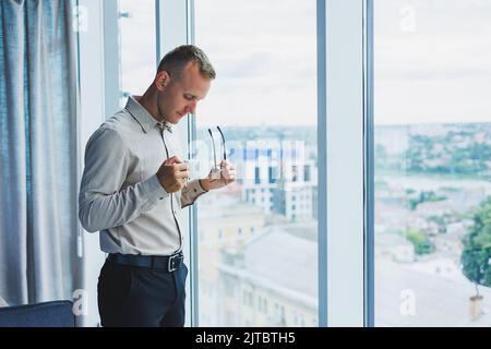 Moderner Bürokaufmann oder Manager, der am Fenster Kaffee trinkt und lächelt. Porträt eines erfolgreichen IT-Ingenieurs-Programmierers, der eine Brille trägt, in der gearbeitet wird Stockfoto