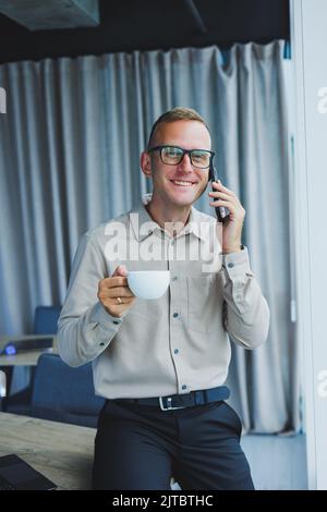 Innenaufnahme des fröhlichen bärtigen männlichen Freiberuflers Arbeit in fernen, verwaltet Finanzen über Smartphone, sitzt am Holztisch, arbeitet mit Dokumentation, hat Stockfoto