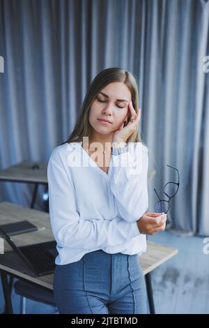 Eine Geschäftsfrau, die mit Kopfschmerzen am Schreibtisch im Büro sitzt. Eine Managerin mit Brille und weißem Hemd leidet an einer Migräne Stockfoto