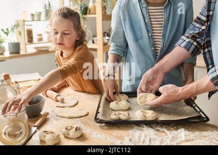 Nahaufnahme der dreiköpfigen Familie, die zum Backen hausgemachte Brötchen auf das Blech legte, kochen sie gemeinsam am Tisch Stockfoto