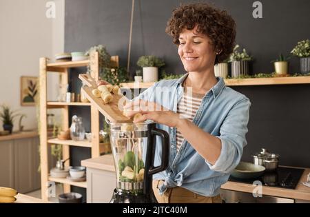 Junge lächelnde Frau, die Bananen in den Mixer gab, während sie in der Küche frischen Smoothie zubereitete Stockfoto