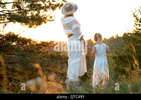 Rückansicht einer wunderbaren Familie in weißen Kleidern, die in der Nähe von Kiefern im Parkwald steht und den Sonnenuntergang im Sommer betrachtet. Stockfoto