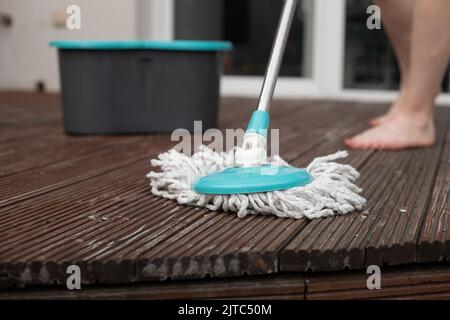 Beschnittenes Foto von Beinen einer jungen Frau, die den braunen Holzboden der Veranda mit einem Spinnmopp in der Nähe eines grauen Plastikeiers putzte. Stockfoto