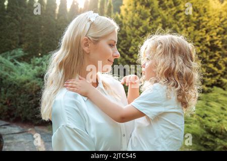 Seitenansicht der Familie, die im Sommer in der Nähe von Kiefern im Park steht. Junge Frau Mutter hält kleines Mädchen in den Armen und trifft auf Augen. Stockfoto