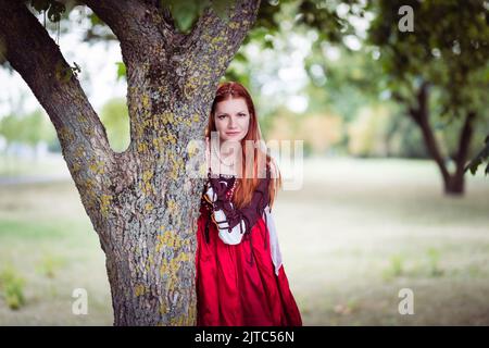 Rothaarige schlanke Mädchen in einem roten Kleid in der venezianischen Mode des 15.. Jahrhunderts posiert in der Nähe eines Baumstamms. Stockfoto