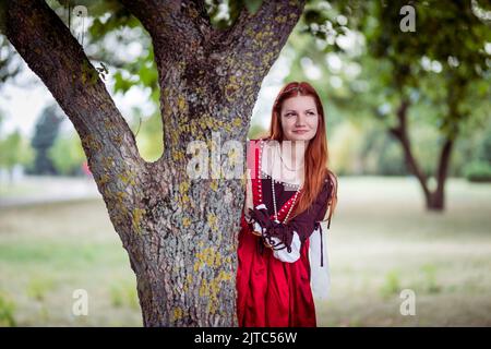 Rothaarige schlanke Mädchen in einem roten Kleid in der venezianischen Mode des 15.. Jahrhunderts posiert in der Nähe eines Baumstamms. Stockfoto