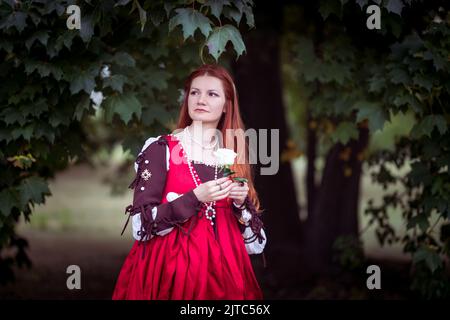 Rothaarige schlanke Mädchen in einem roten Kleid in der venezianischen Mode des 15.. Jahrhunderts posiert unter einem großen Ahornbaum. Stockfoto