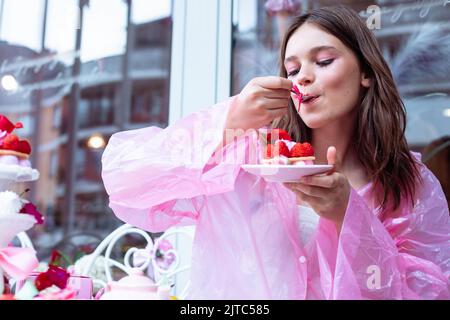 Hübsches und ruhiges Teenager-Mädchen in rosa Regenmantel, das fröhlich ist und süße Himbeer-Dessertkuchen im Café im Freien in der Nähe des Fensters verkostet Stockfoto