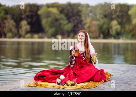Rothaarige schlanke Mädchen in einem roten Kleid in der venezianischen Mode des 15.. Jahrhunderts hält ein paar Trauben und einen Becher Getränk in den Händen Stockfoto