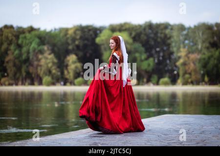 Rothaarige schlanke Mädchen in einem roten Kleid in der venezianischen Mode des 15.. Jahrhunderts. Das Mädchen dreht sich auf dem Pier des Sees, ihr Rock flattert Stockfoto