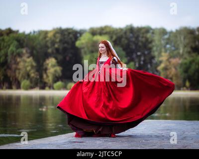 Rothaarige schlanke Mädchen in einem roten Kleid in der venezianischen Mode des 15.. Jahrhunderts. Das Mädchen dreht sich auf dem Pier des Sees, ihr Rock flattert Stockfoto