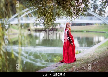 Rothaarige schlanke Mädchen in einem roten Kleid in der venezianischen Mode des 15.. Jahrhunderts posiert vor einem kleinen Teich, voller Länge Porträt Stockfoto