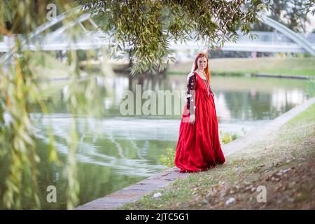 Rothaarige schlanke Mädchen in einem roten Kleid in der venezianischen Mode des 15.. Jahrhunderts posiert vor einem kleinen Teich, voller Länge Porträt Stockfoto