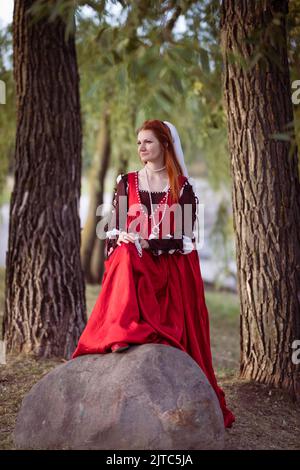 Rothaarige schlanke Mädchen in einem roten Kleid in der venezianischen Mode des 15.. Jahrhunderts posiert in der Nähe eines großen Steins. Stockfoto