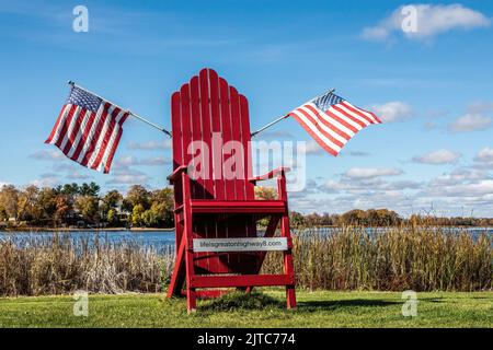 Großer roter Adirondack-Stuhl mit zwei amerikanischen Flaggen vor dem North Center Lake in Center City, Minnesota, USA. 8. Stockfoto