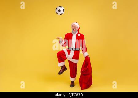 In voller Länge älterer Mann mit grauem Bart trägt weihnachtsmann Kostüm spielt mit Fußball, hält rote Tasche Taschen mit Neujahr Geschenke. Innenaufnahme des Studios isoliert auf gelbem Hintergrund. Stockfoto